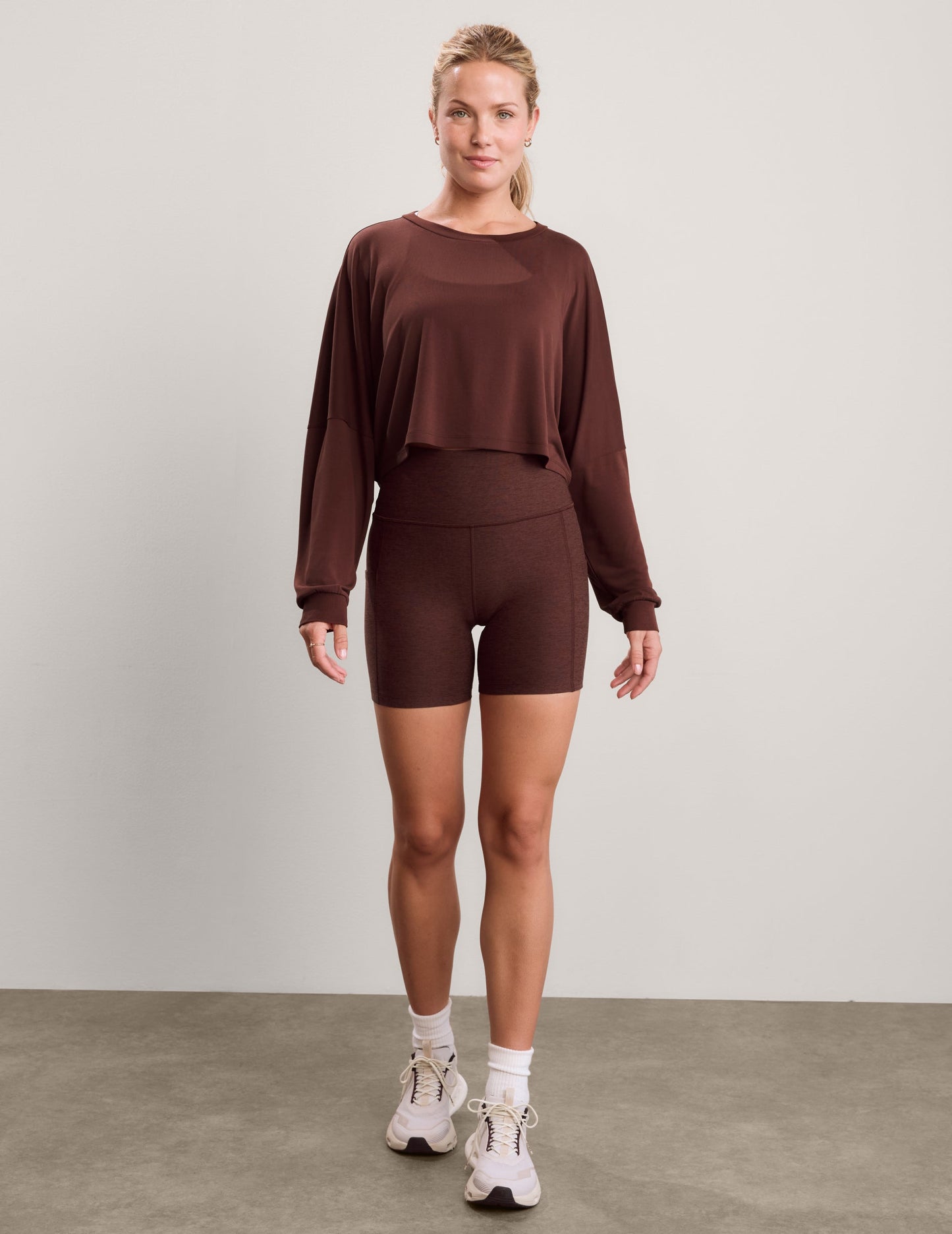 Woman wearing a brown long sleeve top and brown biker shorts against a plain background.