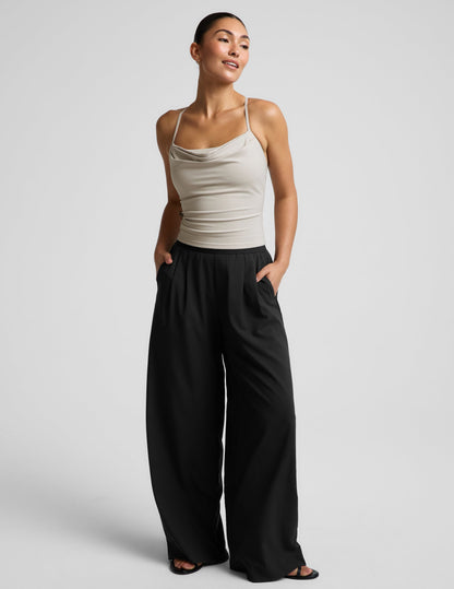 Woman wearing a beige cowl neck tank top and black trousers against a plain background.