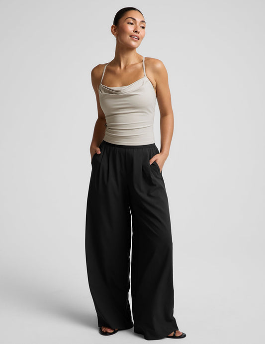 Woman wearing a beige cowl neck tank top and black trousers against a plain background.