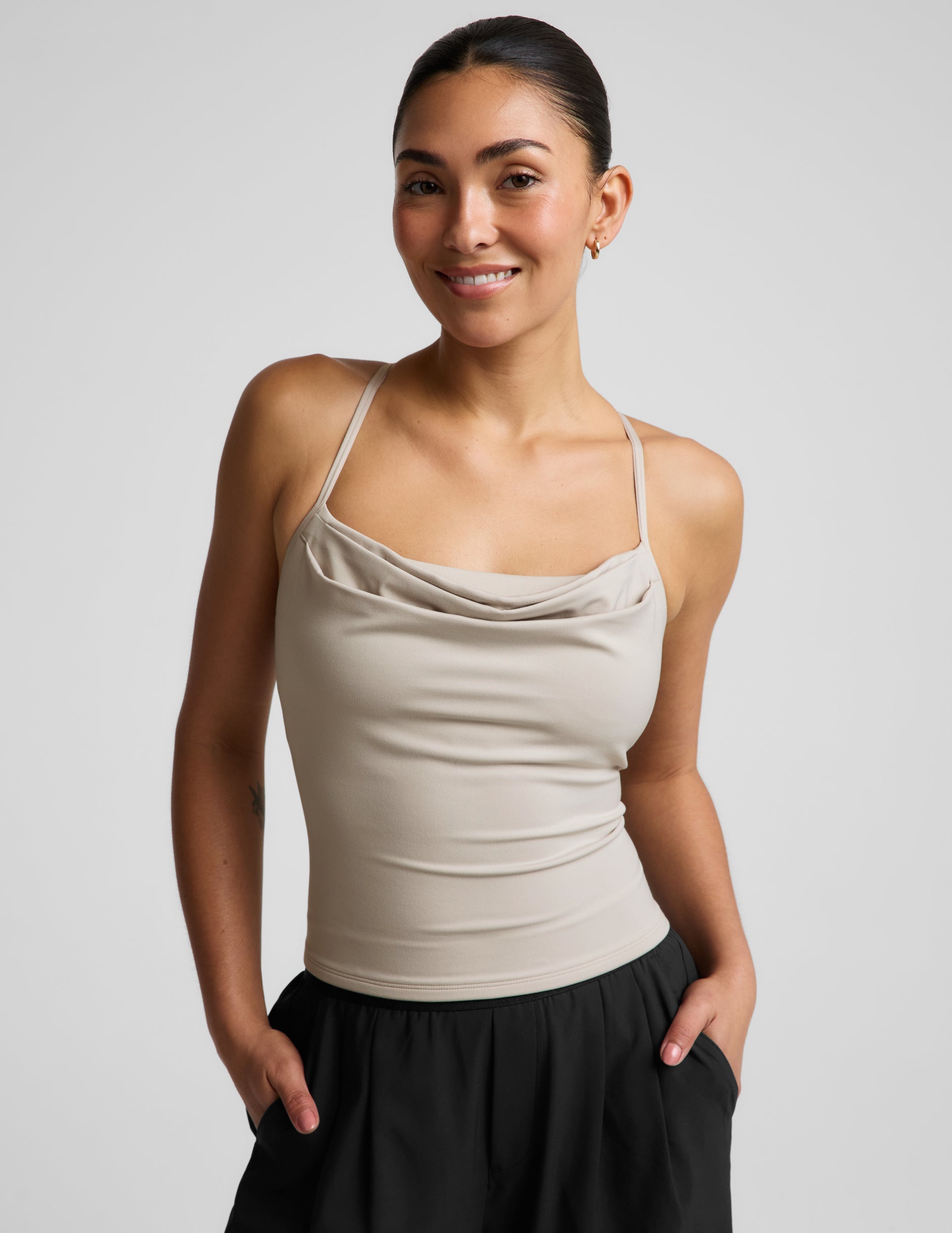 Woman wearing a beige cowl neck tank top and black trousers against a plain background.