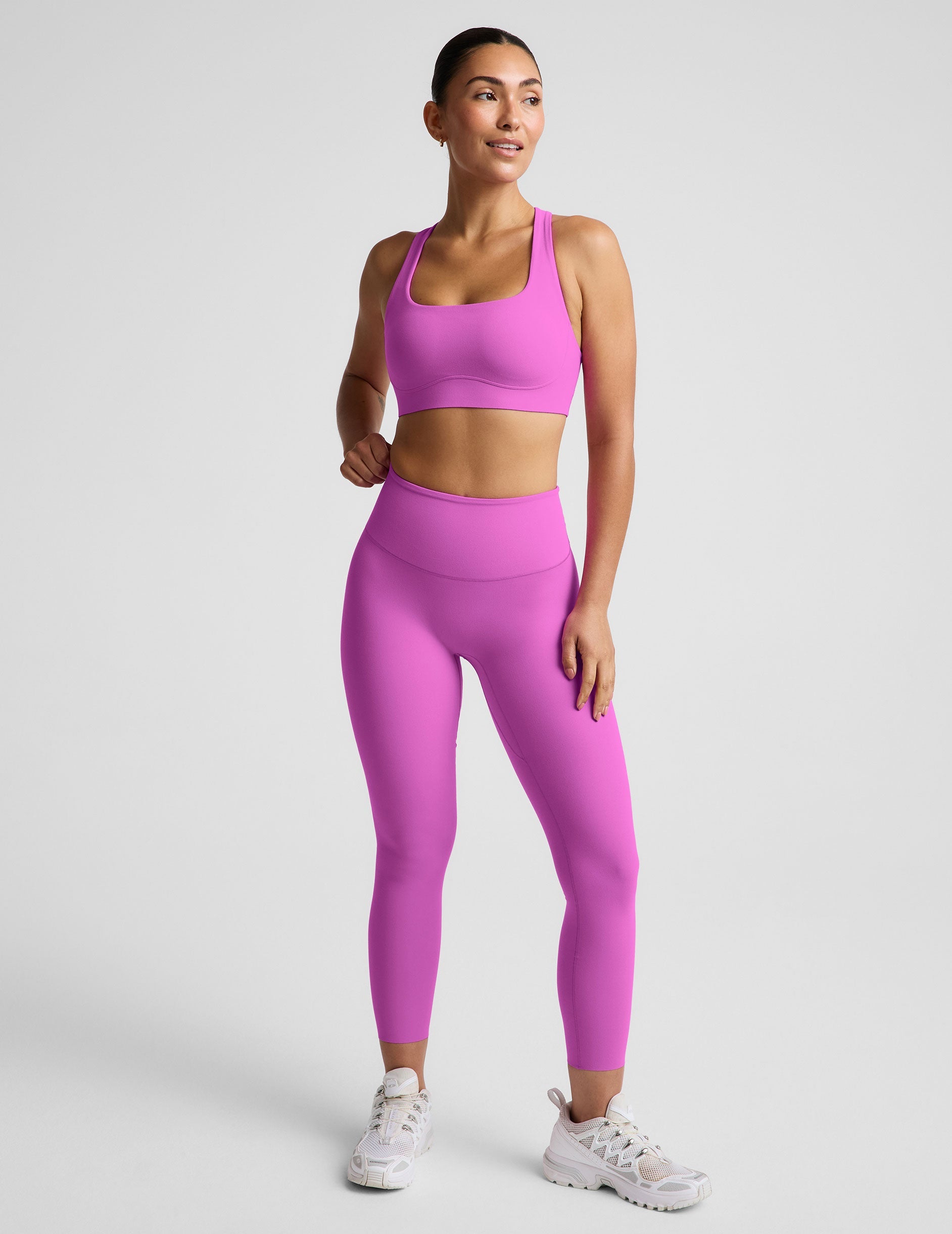Woman wearing a purple sports bra and purple midi length leggings against a plain studio background.