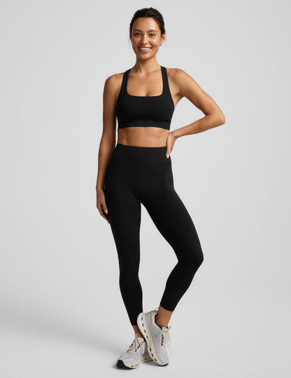 A woman in a black sports bra and black midi length leggings against a plain background.