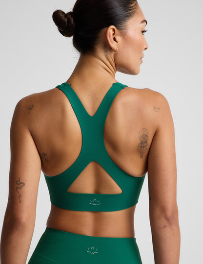 A woman shown from the back wearing a dark green athletic sports bra and matching high-waisted bottoms against a light gray studio background.