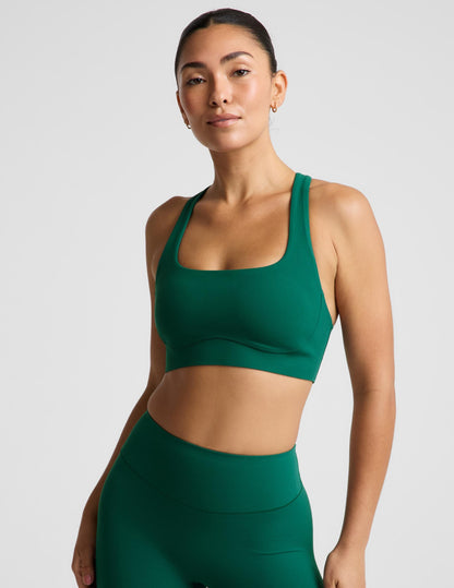 A woman modeling a dark green athletic sports bra and matching high-waisted leggings against a light gray studio background.