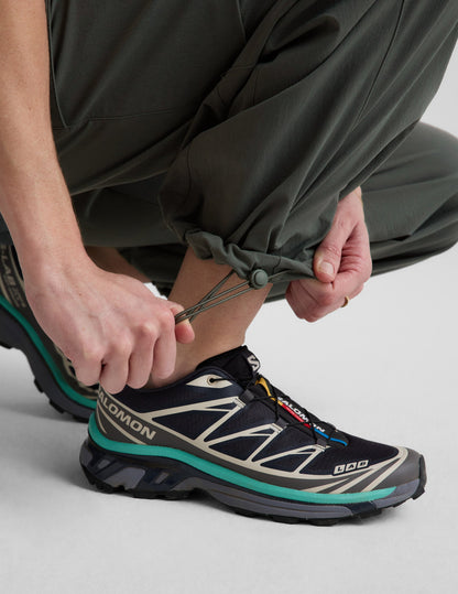 Close-up of woman adjusting the bungee hem on the Dark Olive Peak Pant™ paired with black and teal sneakers.