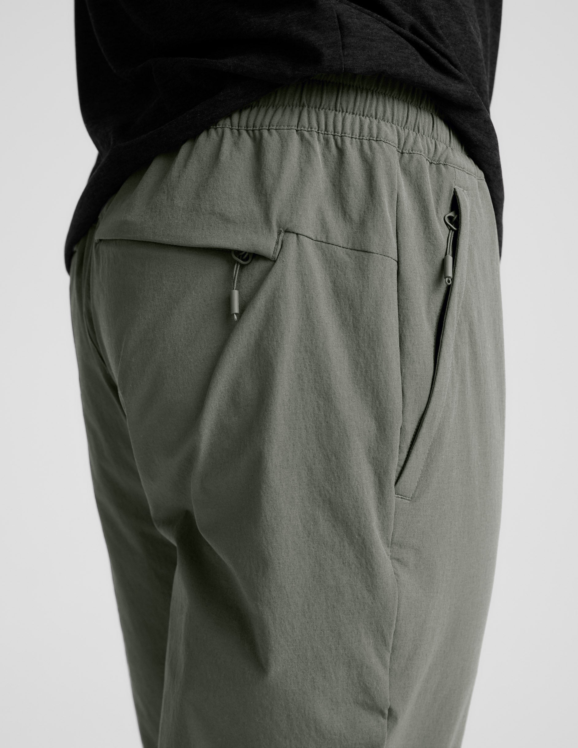 Side view of olive Men's All Terrain Pant showing gathered elastic waistband and side and back zipper pockets with pulls.