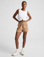 Woman wearing tan Peak™ 3" Short with a knotted white tank top, featuring a drawstring waist and side zip cargo pockets.