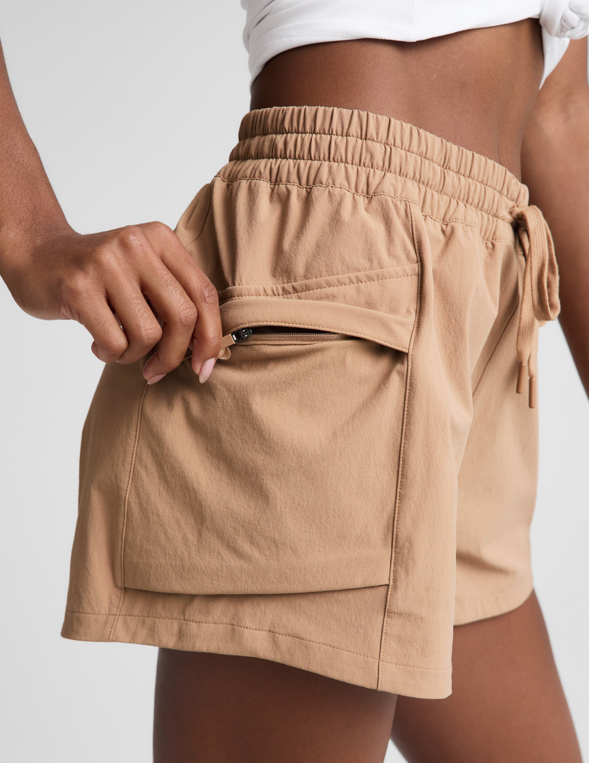 Woman wearing Tan Peak 3" Short™ with a white top, showing the side patch cargo pocket and zipper closure.