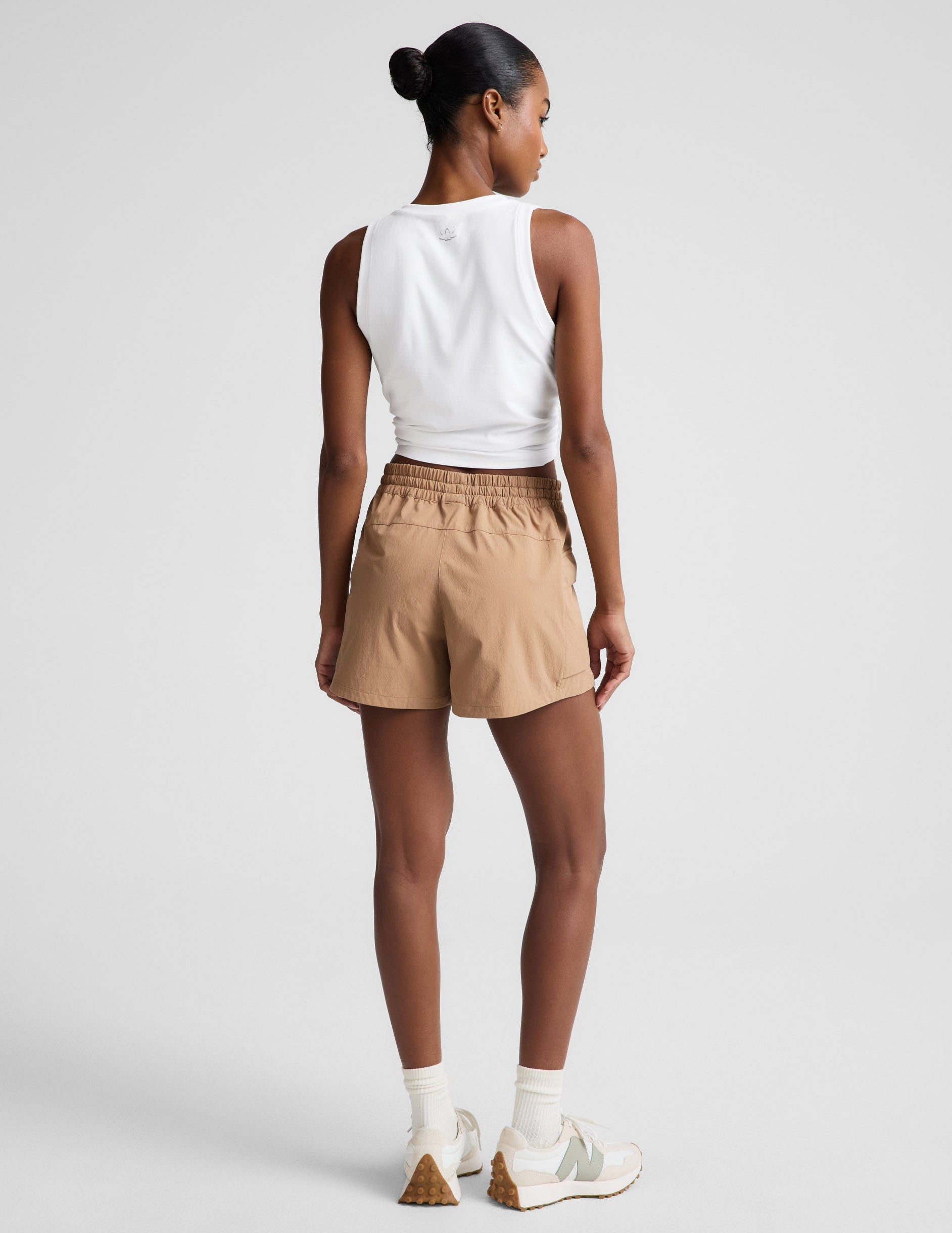Woman wearing tan Peak 3" Short with a white cropped tank top, rear view showing the gathered elastic waistband.