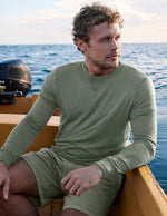 Man wearing a green long-sleeve shirt and shorts sitting on a boat with water in the background.