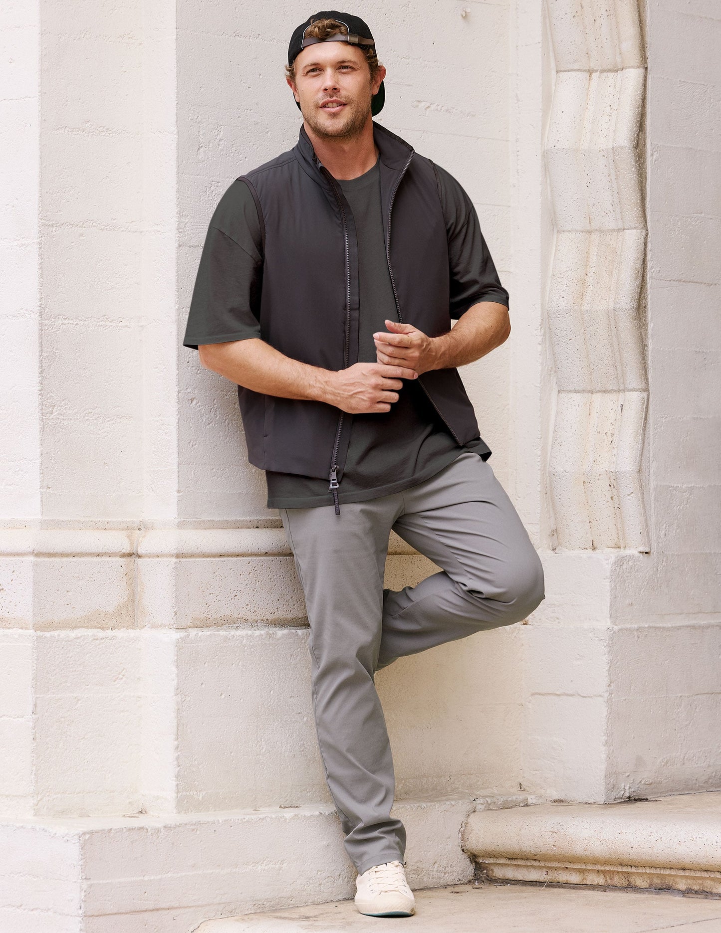 Man in a black vest and grey Element Men's Chino Trouser leans against a white stone wall, showcasing the slim fit.