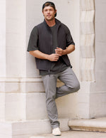 Man in a black vest and grey Element Men's Chino Trouser leans against a white stone wall, showcasing the slim fit.