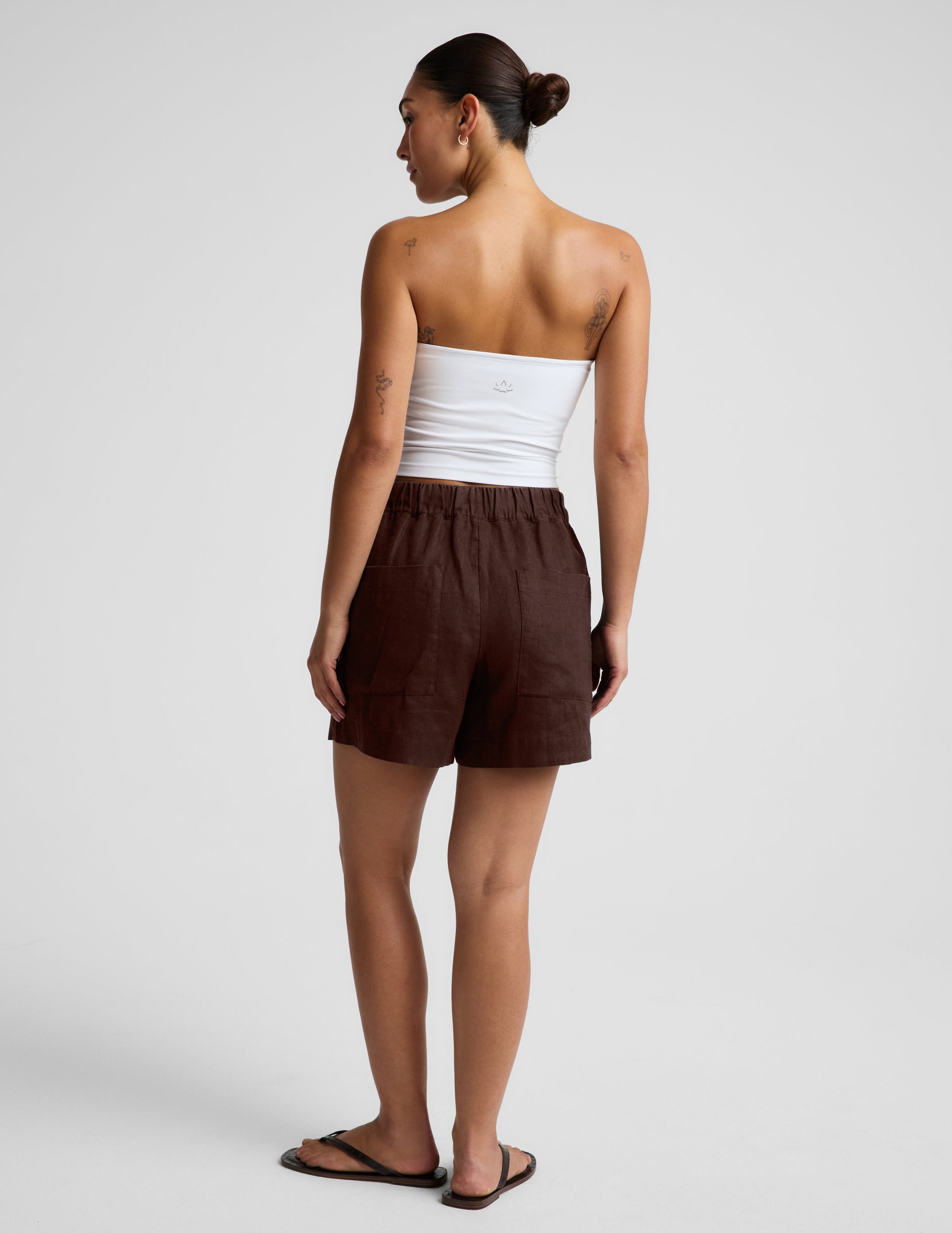 Woman wearing a white strapless tank and brown linen shorts against a plain background.