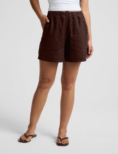 Woman wearing a white strapless tank and brown linen shorts against a plain background.
