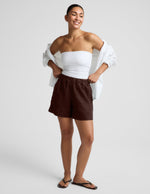 Woman wearing a white strapless tank and brown linen shorts against a plain background.