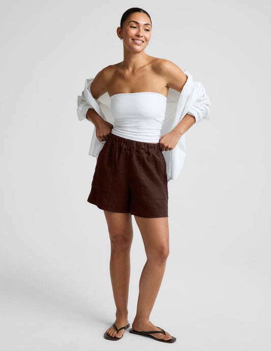 Woman wearing a white strapless tank and brown linen shorts against a plain background.